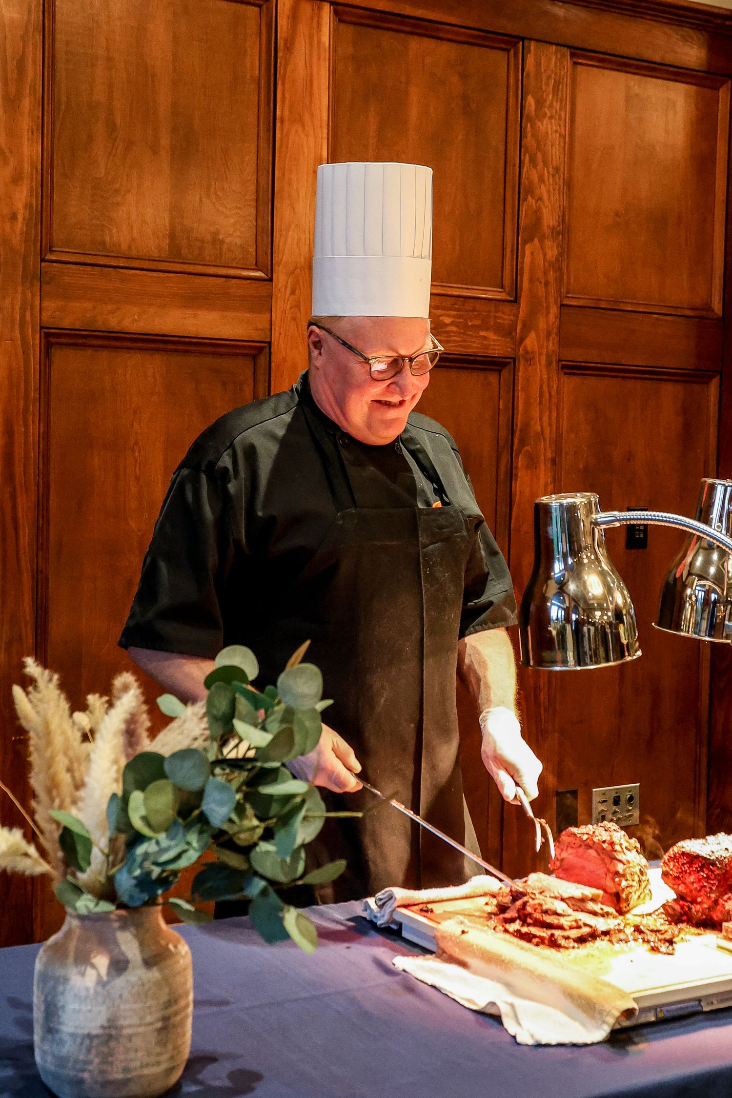Chef in white hat carves roasted meat at an elegant wood-paneled banquet station.