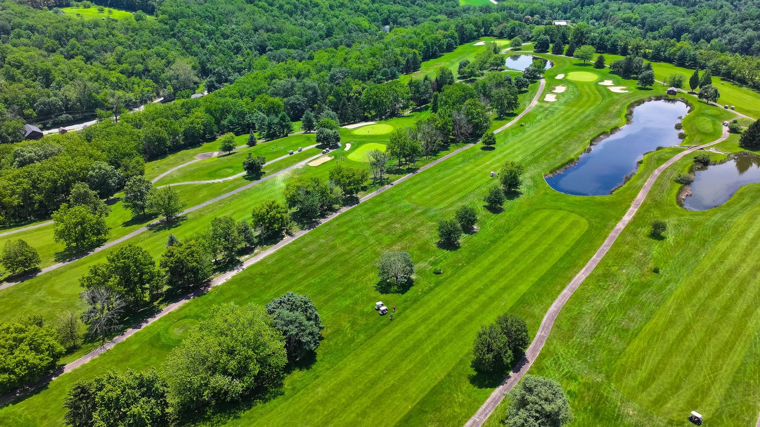 Aerial panoramic view of a lush green golf course with rolling fairways, sand bunkers, and surrounding forest landscape.