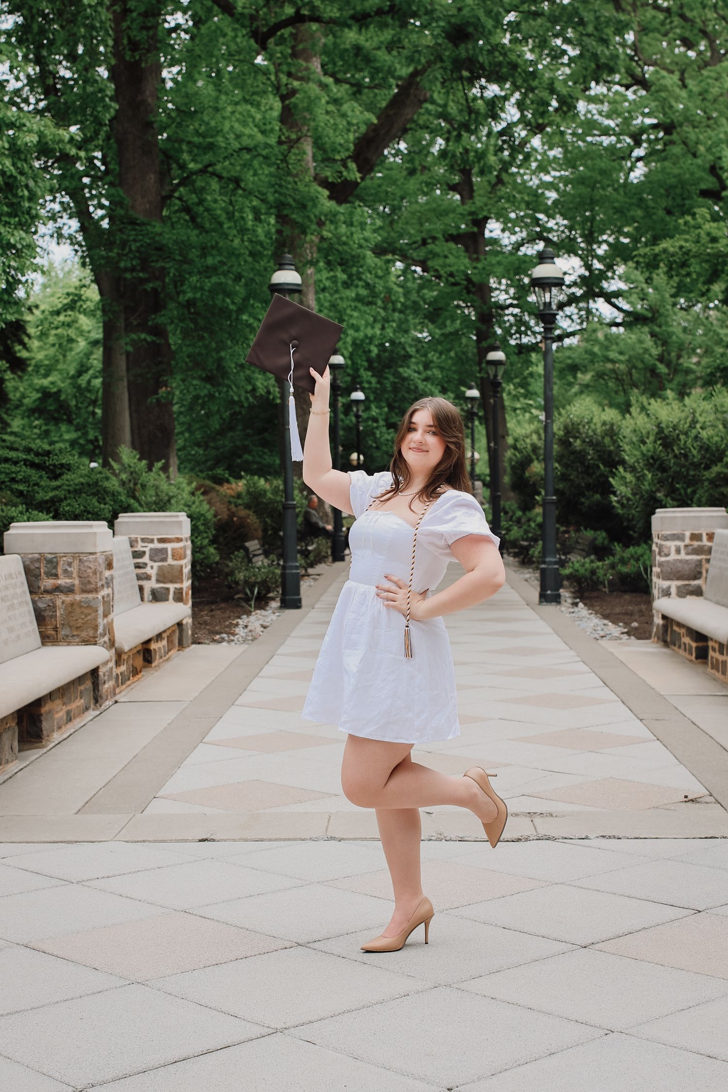 A person in white dress performs graceful dance movements on a stone patio surrounded by lush green trees.