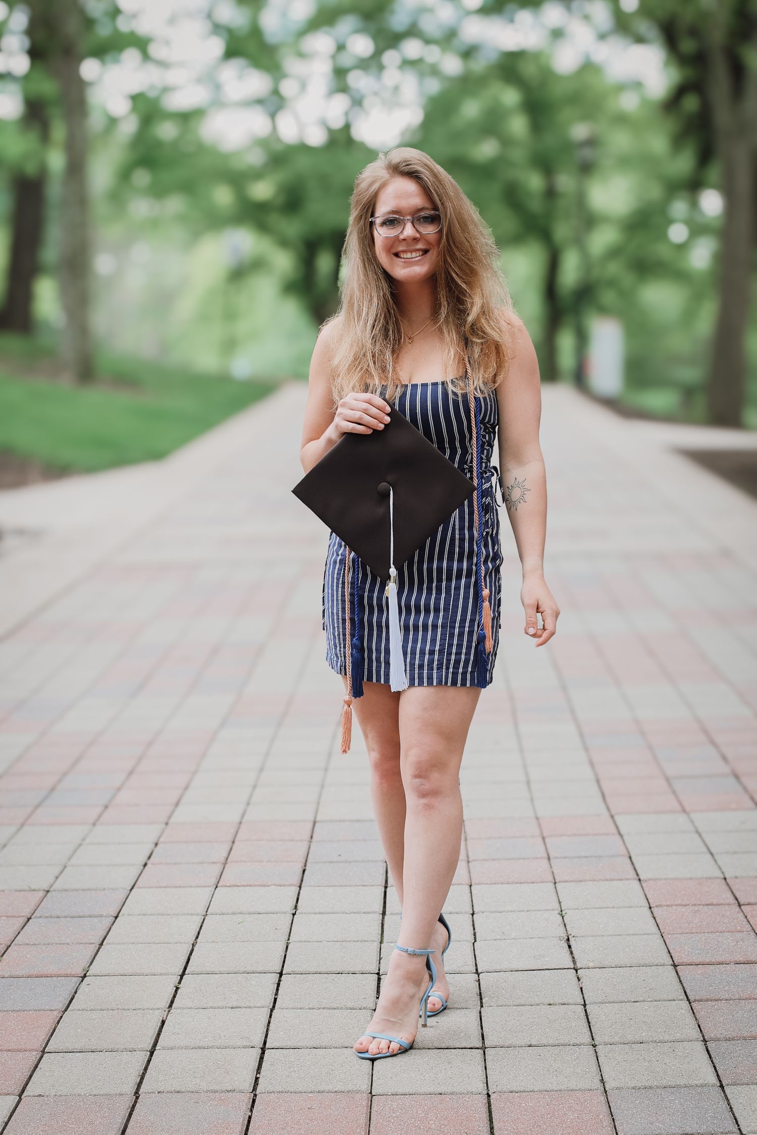 Graduate in striped dress poses playfully on brick pathway surrounded by green trees during photo session.