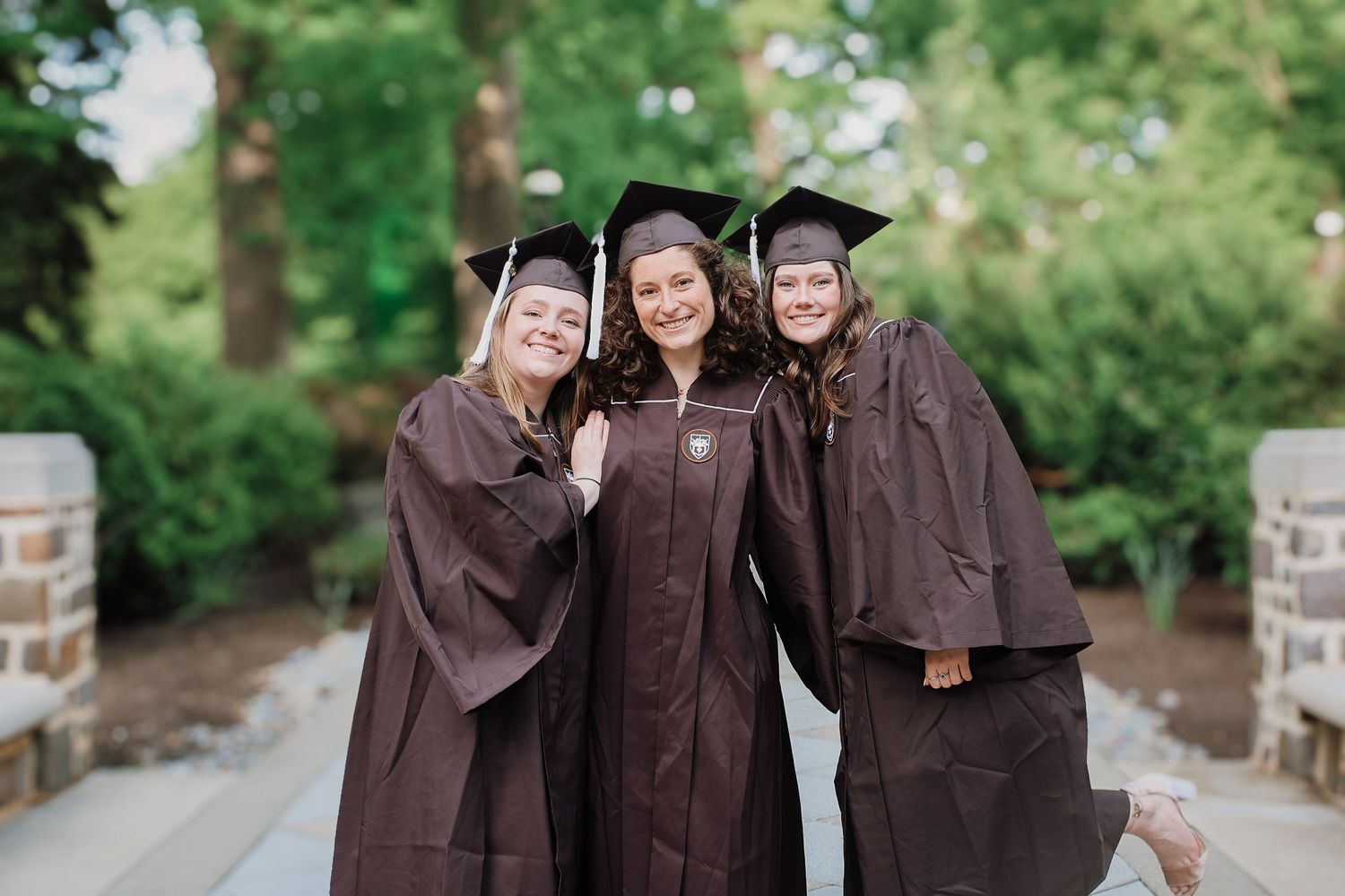 A group in brown graduation gowns poses together outdoors on a sunny day near brick columns.