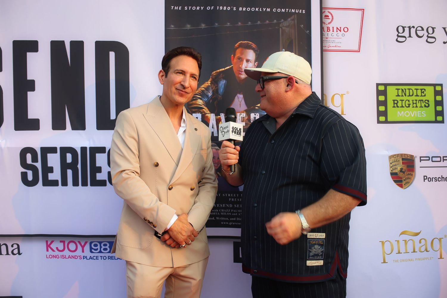 Two people in formal attire shake hands at a media event with sponsor logos visible on the backdrop.