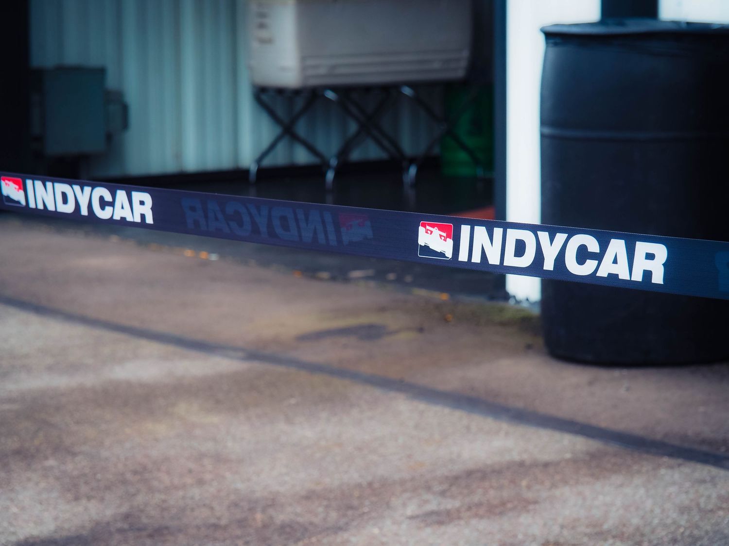 An IndyCar racing banner hangs above a pit lane entrance at a motorsports track during a race event.