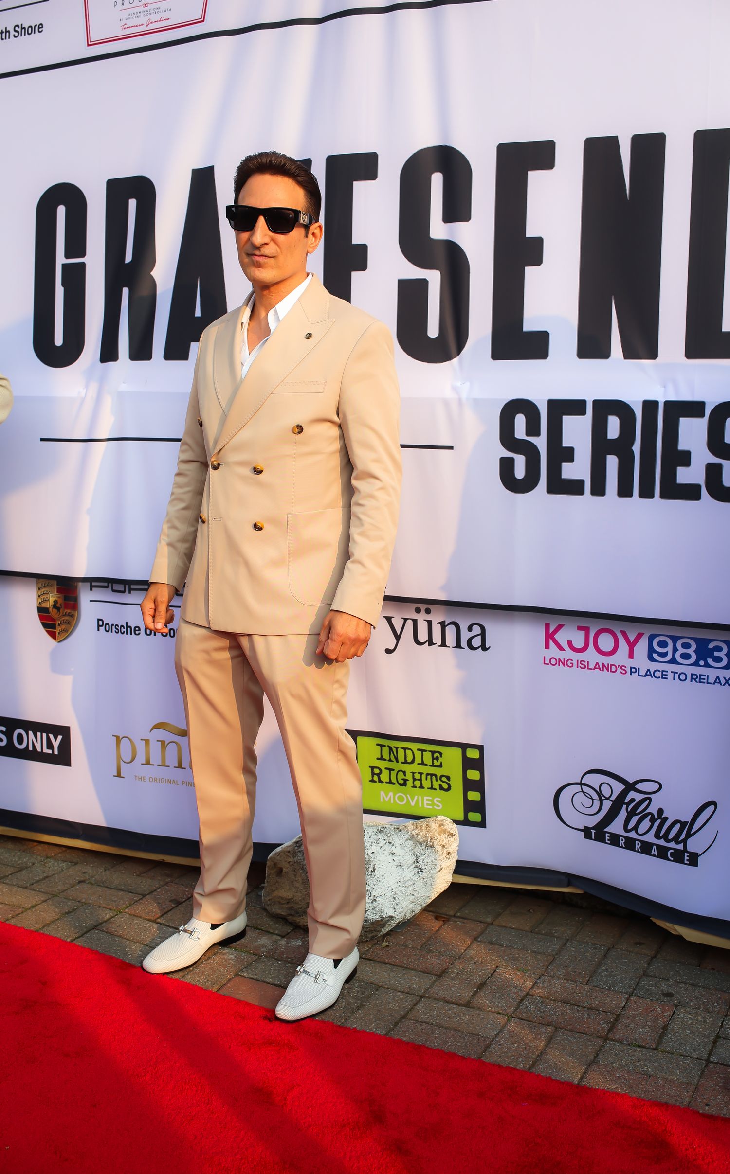 A person wearing a cream-colored suit and sunglasses poses on a red carpet against a branded step-and-repeat backdrop.