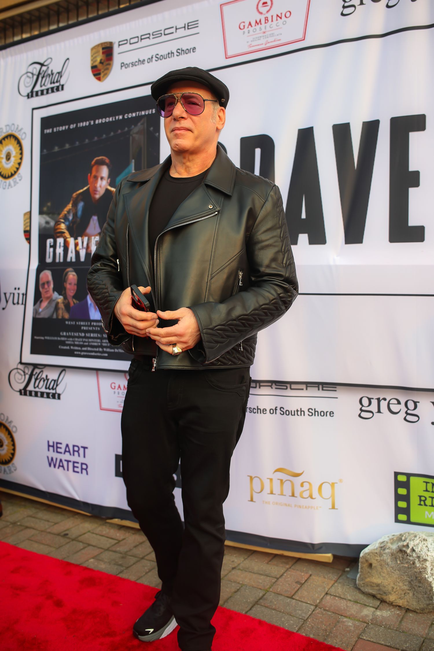 A person in black leather jacket and sunglasses poses on a red carpet with event sponsor logos visible in the background.