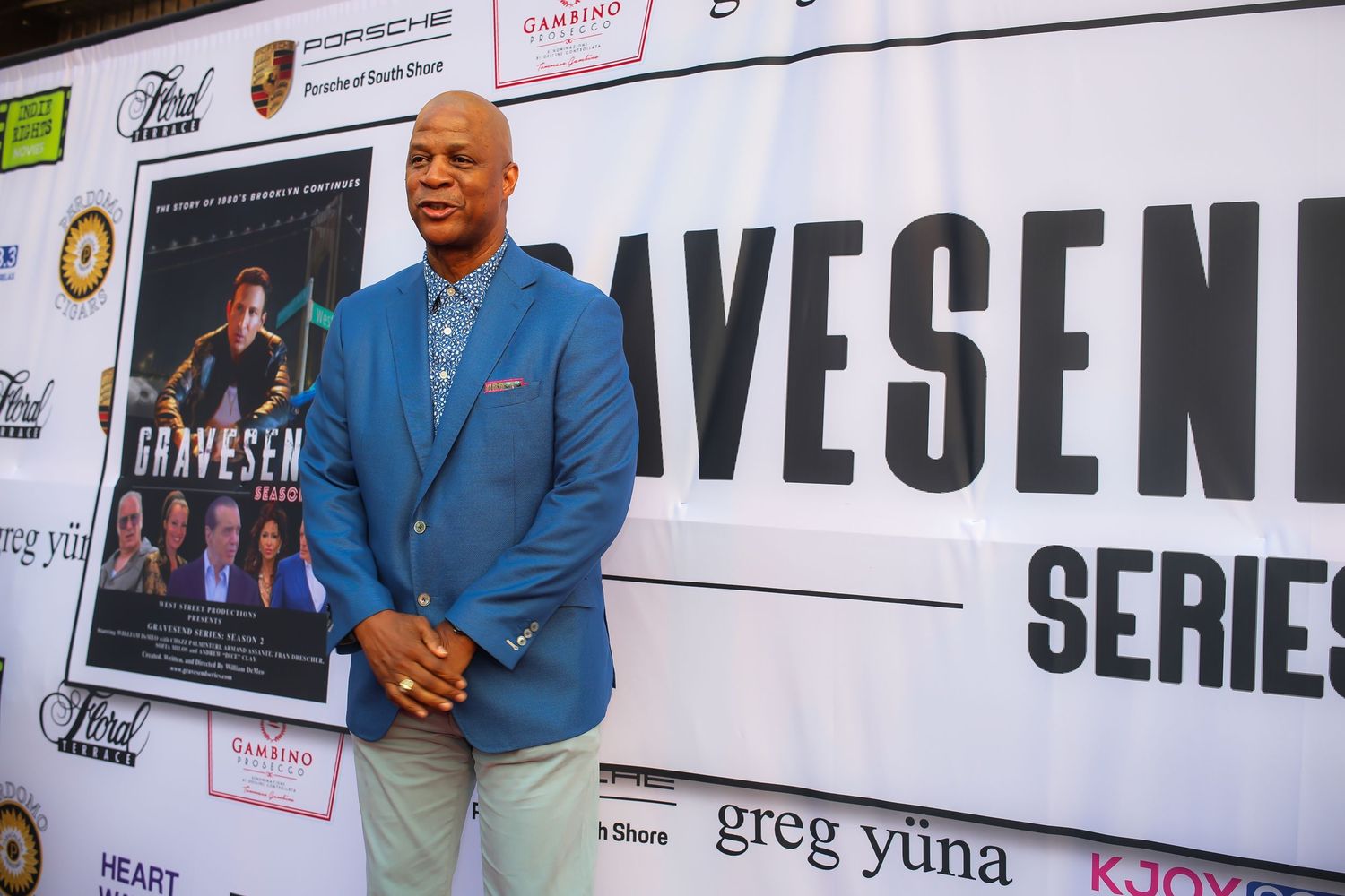 A person in a light blue blazer and khaki pants poses against a backdrop with text at a promotional event.