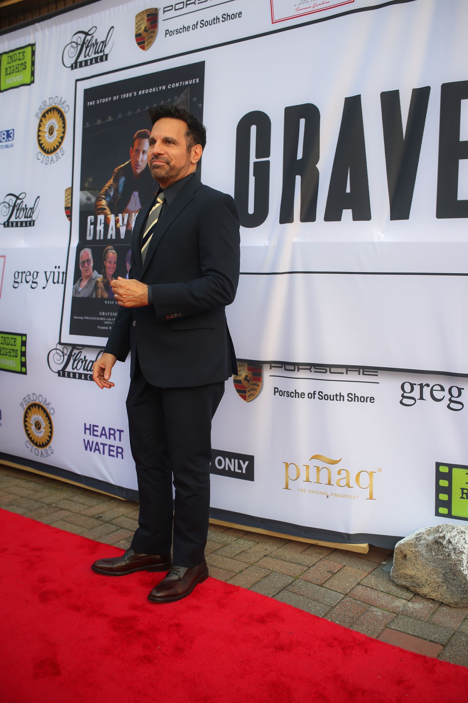 A red carpet event backdrop with sponsor logos and a person standing in formal attire at a media wall.