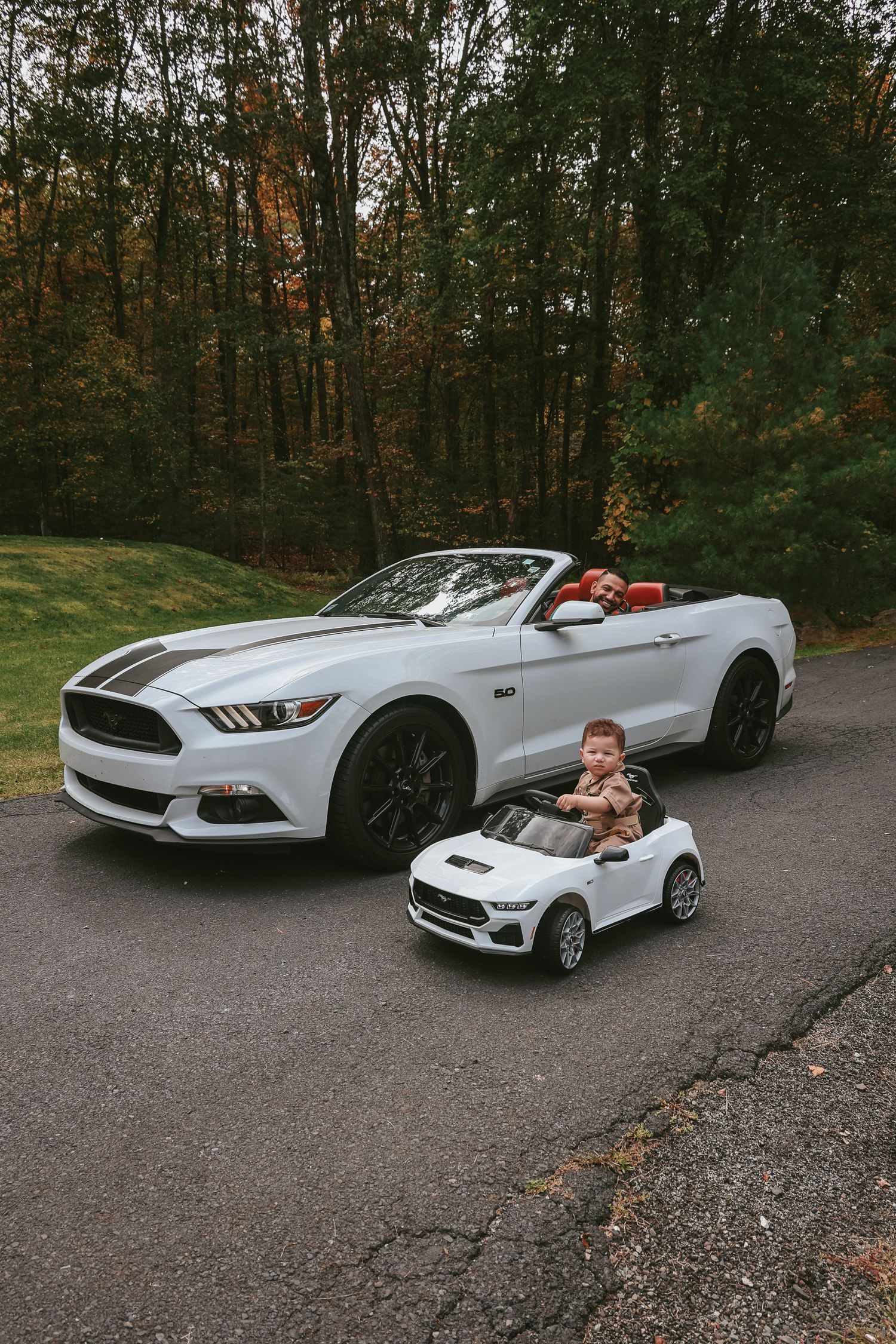 White Ford Mustang convertible parked on a wooded road during autumn with fall foliage in the background.