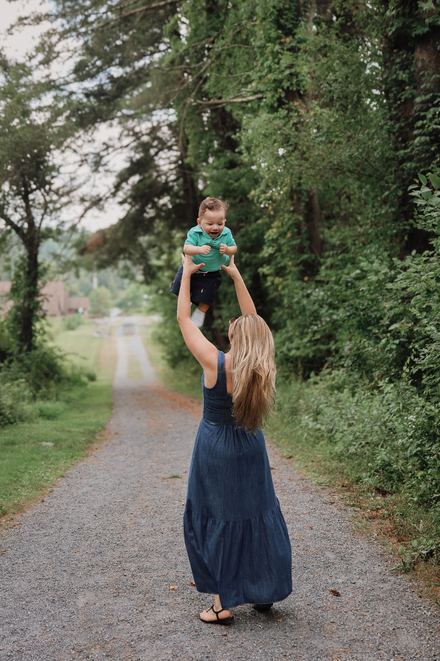 A person in a navy blue dress walks down a tree-lined gravel path on a summer day.