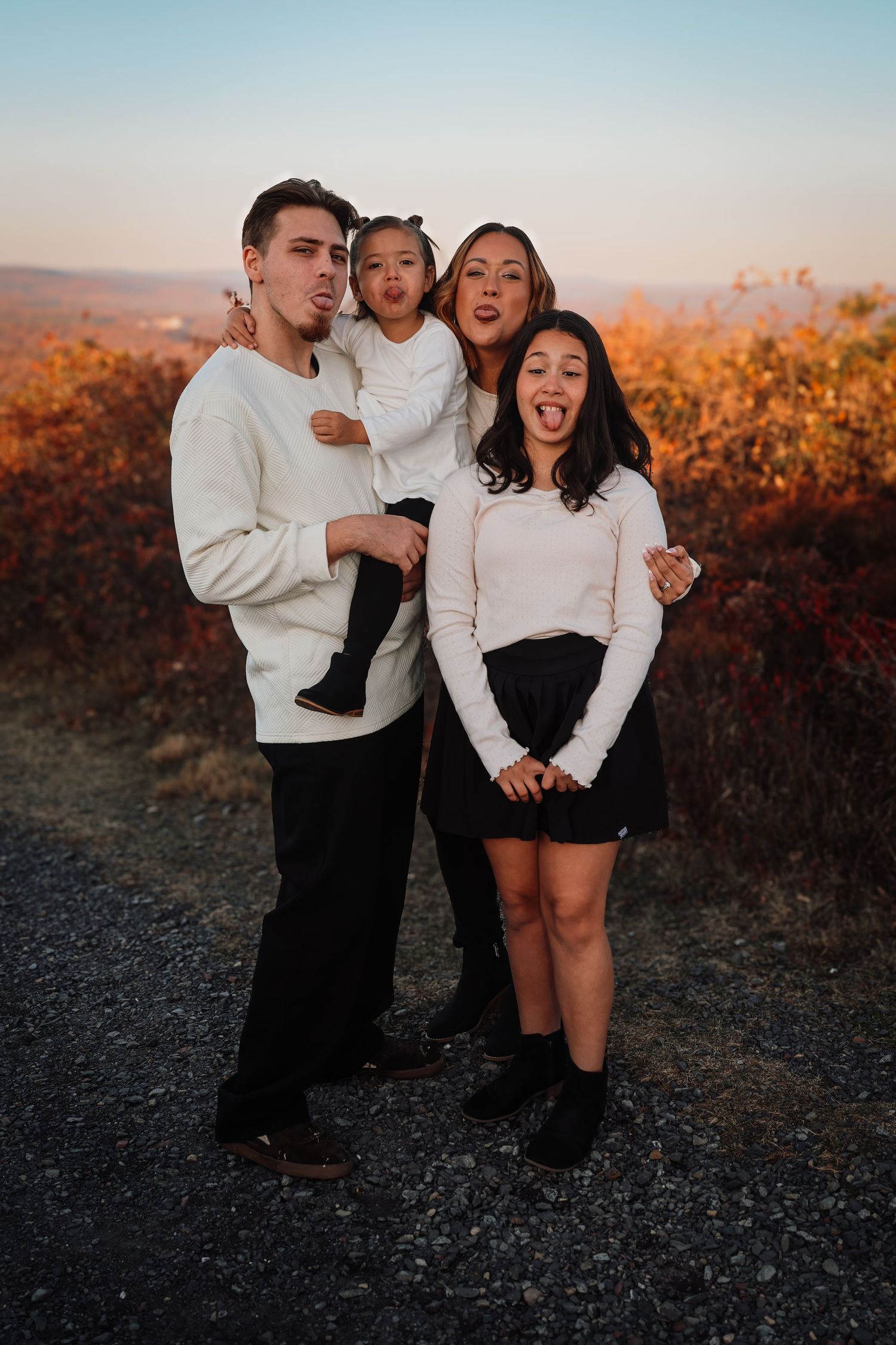 A family in matching white outfits poses playfully at sunset against autumn foliage.