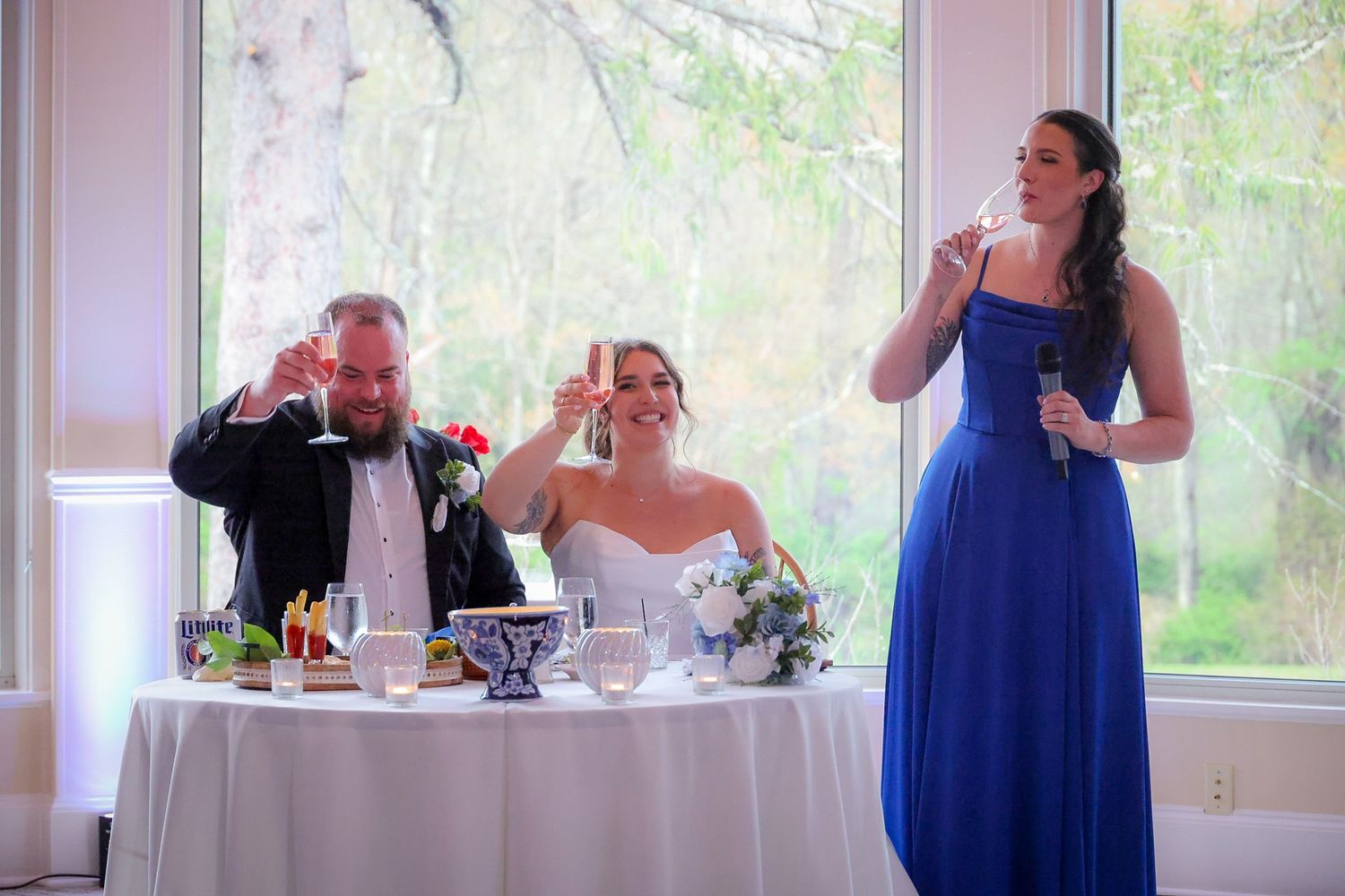 Bride and groom toast with bridesmaid in blue dress giving speech at elegant wedding reception.