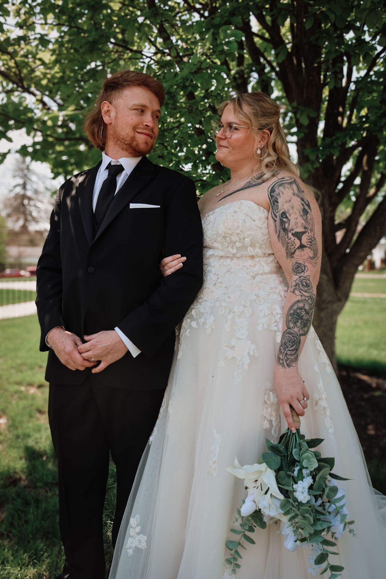 Bride and groom pose outdoors under green trees, she wears a floral gown with tattoos, he wears a black tuxedo.