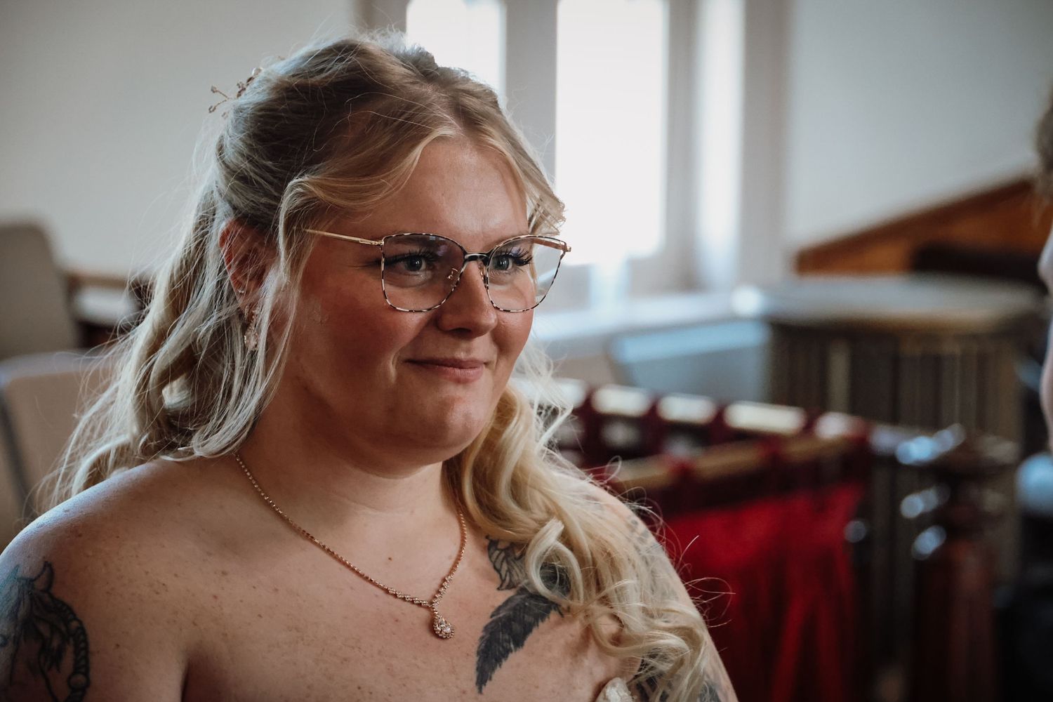Smiling woman with glasses and tattoos wearing a necklace, posed indoors in a candid portrait photo.