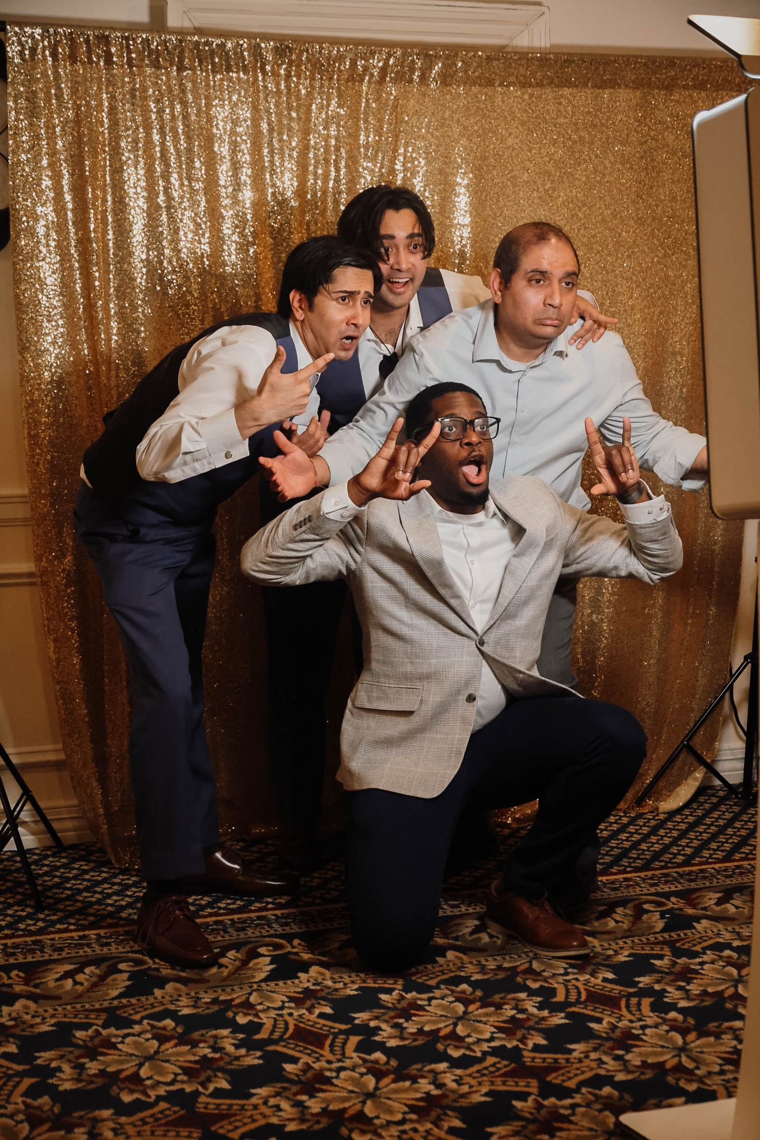 Four men posing playfully in front of a gold sequin backdrop at a formal event photo booth.
