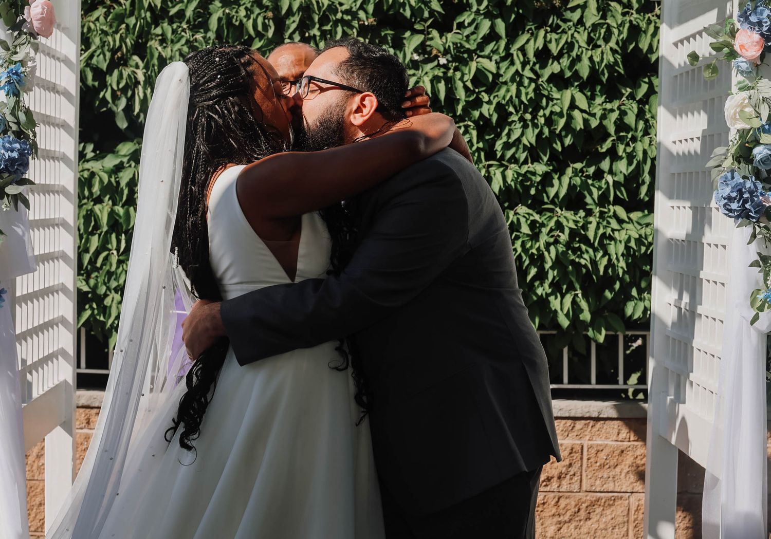 Bride and groom sharing a romantic embrace at outdoor wedding ceremony with white arbor and blue hydrangeas.