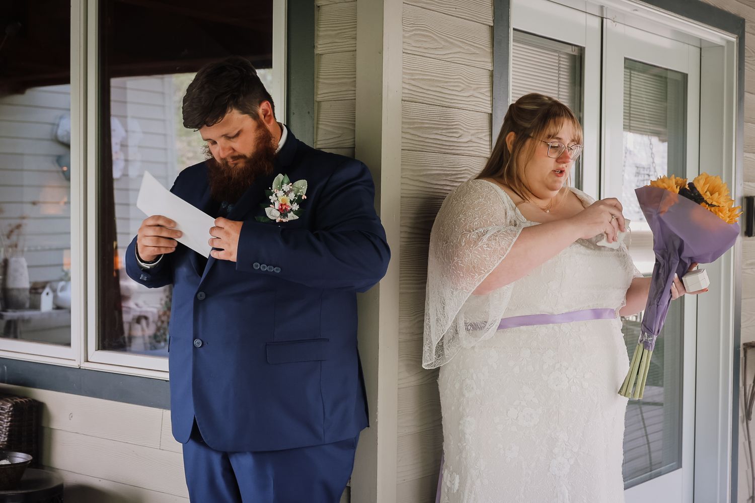Bride holding sunflowers and groom in blue suit reading vows on porch before wedding ceremony.