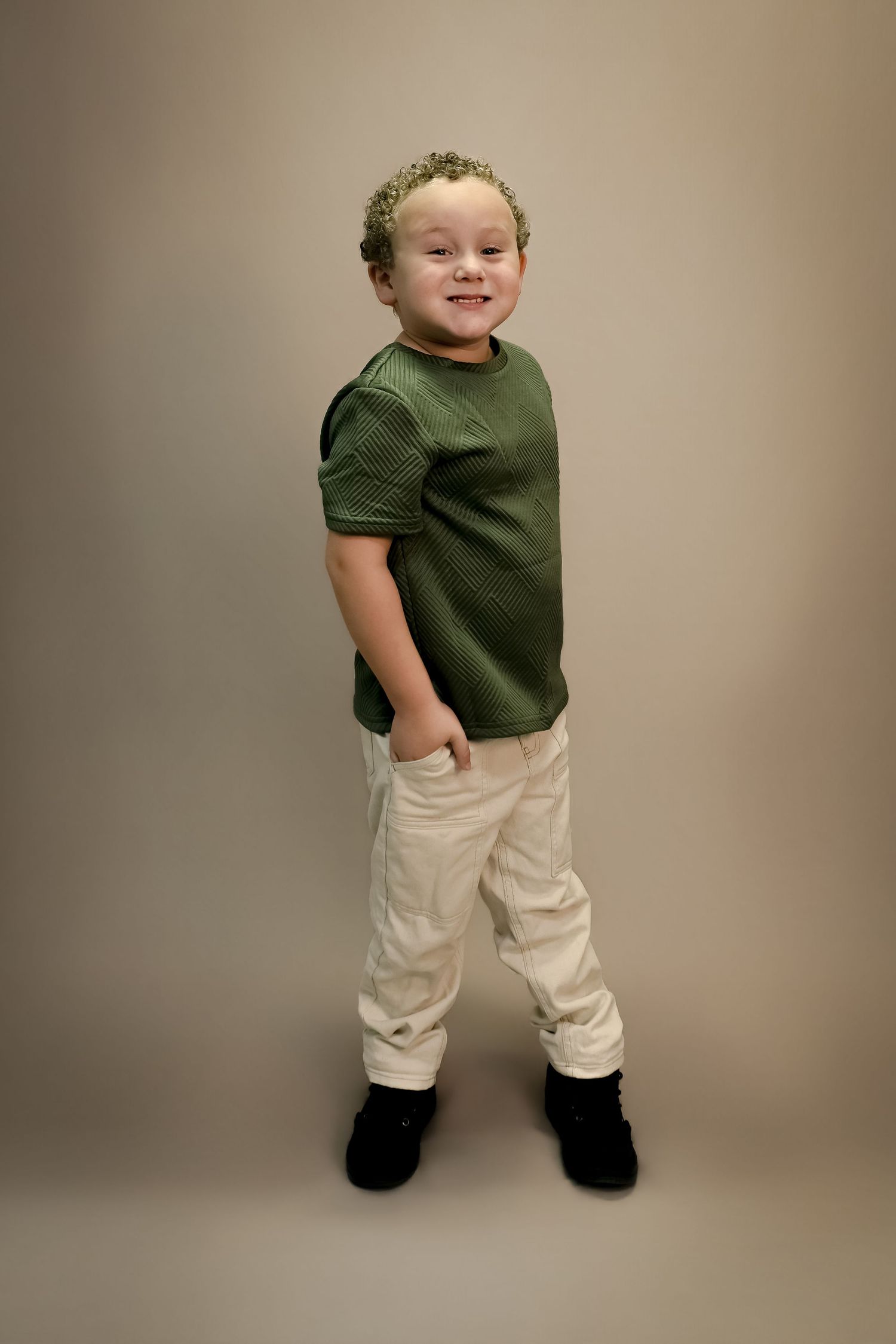 A smiling young boy with curly hair wearing a green t-shirt, cream pants, and black shoes poses confidently.