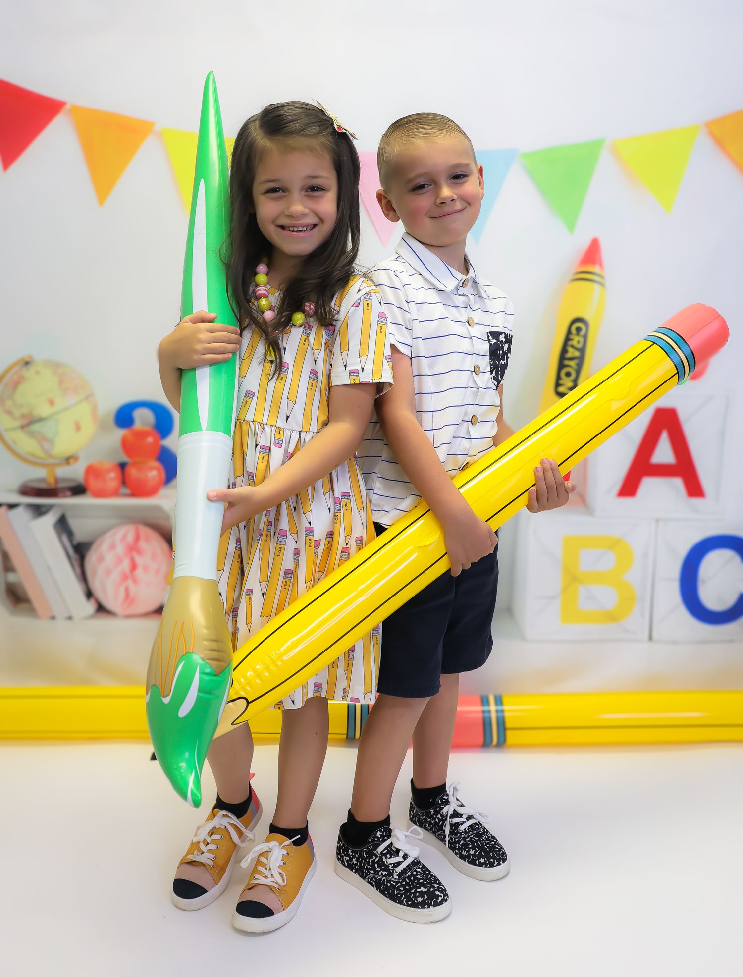 Two children pose with oversized inflatable pencil and crayon toys in front of colorful back-to-school decorations.