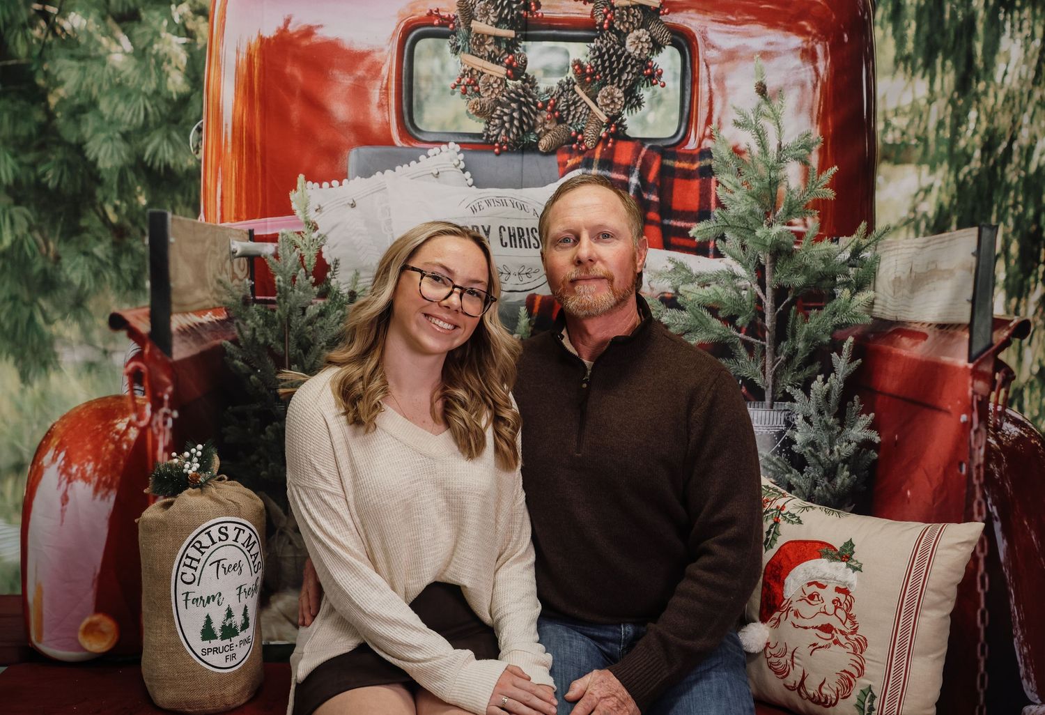 A couple smiles together in front of a festive Christmas truck backdrop with holiday decorations and pillows.
