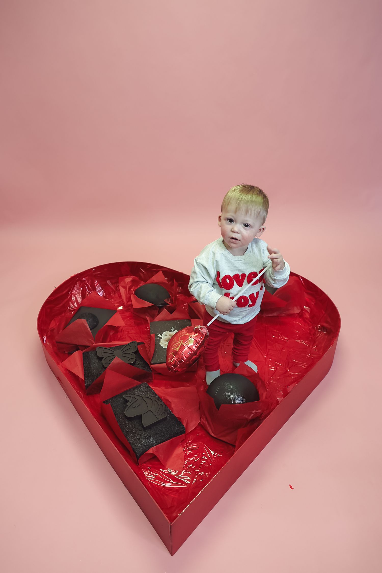 Toddler in love shirt sits inside giant red heart-shaped Valentine's Day chocolate box on pink background.