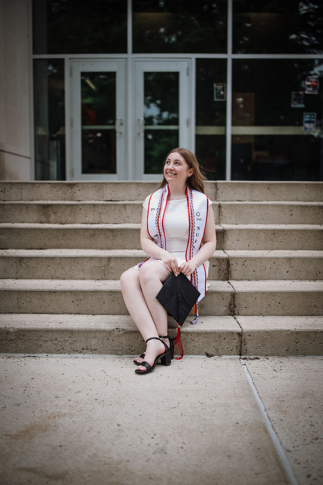 Young woman in graduation attire sits on concrete steps holding cap, wearing white dress and honor stole.