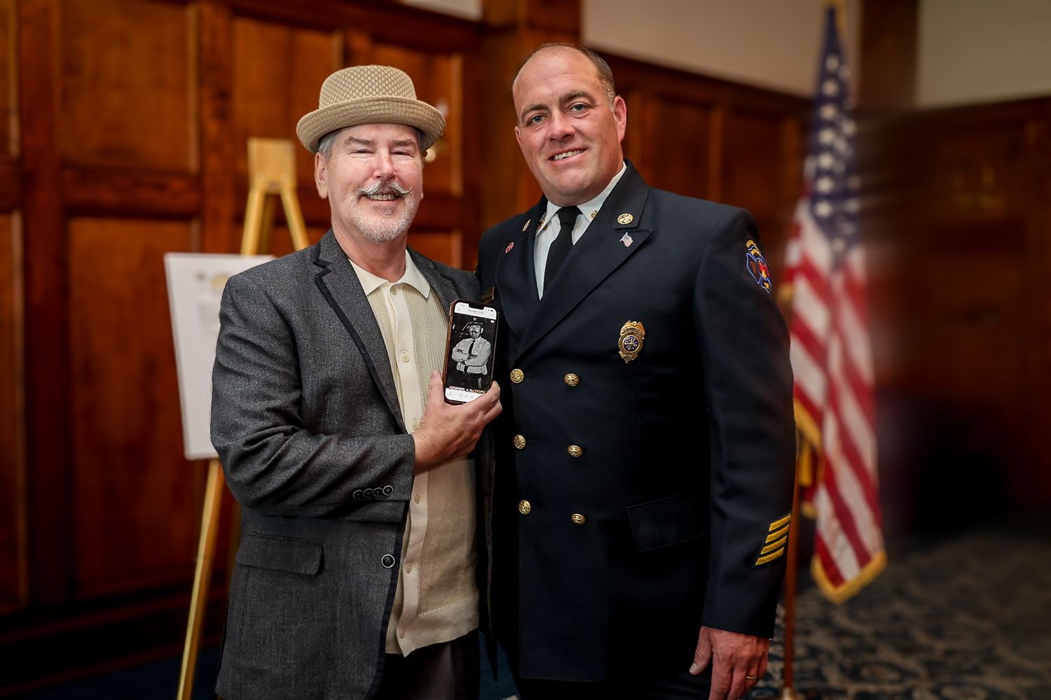Two men pose together, one in a suit holding a badge, the other in a fire department uniform, with a US flag in the background.