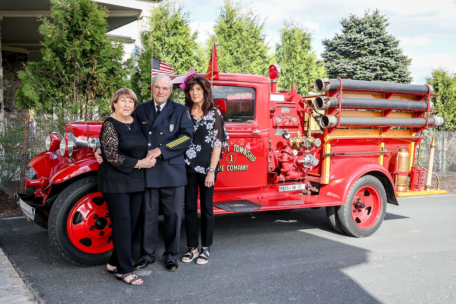 Three people pose proudly beside a vintage red fire truck outdoors, with lush greenery in the background.