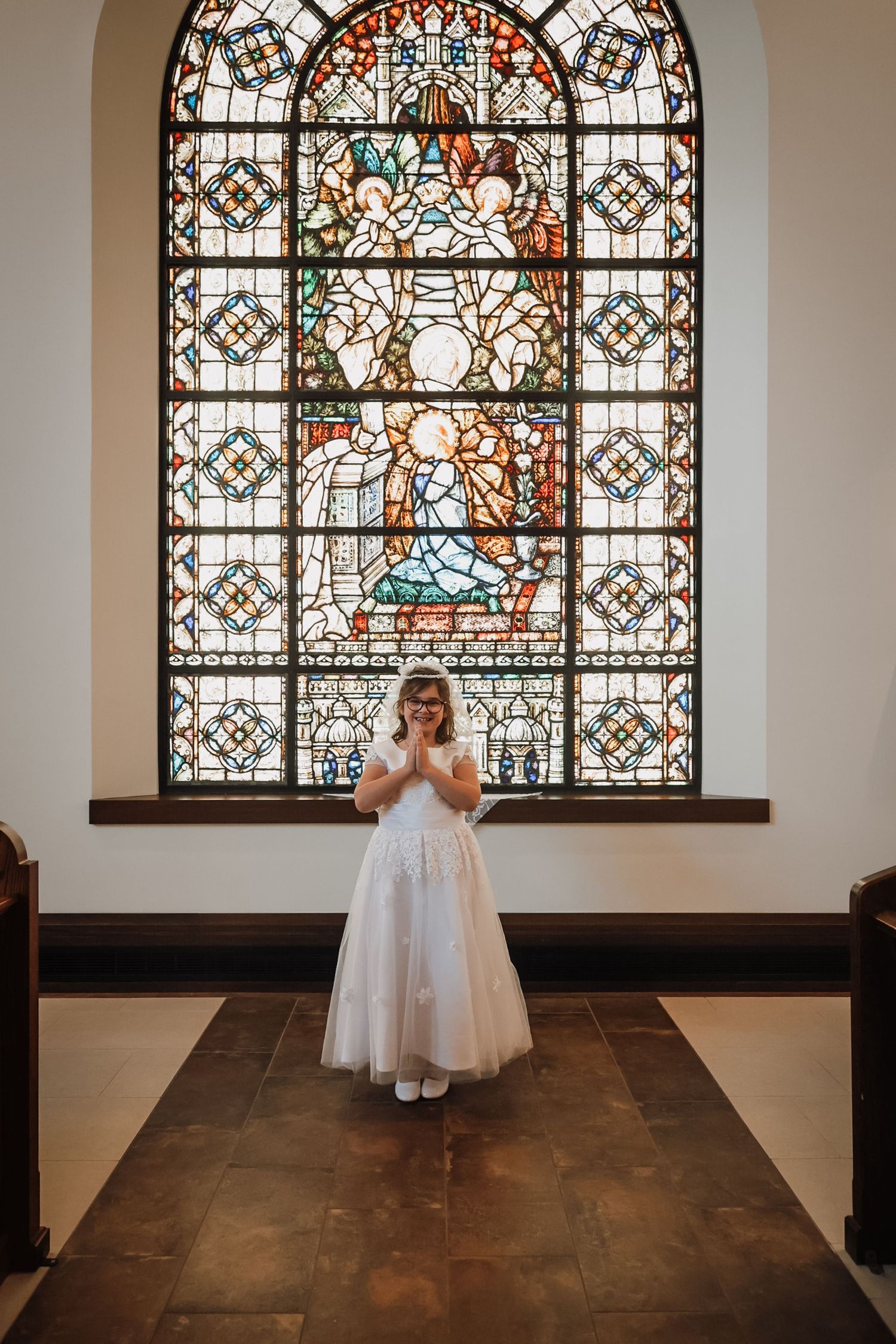 Young girl in white dress stands before ornate stained glass window in church during First Communion ceremony.