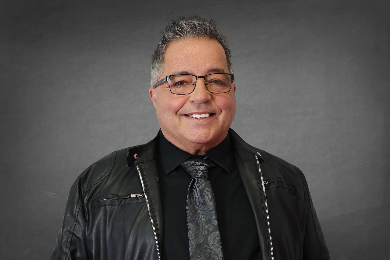 A person wearing glasses and a black leather jacket poses against a dark gray background.