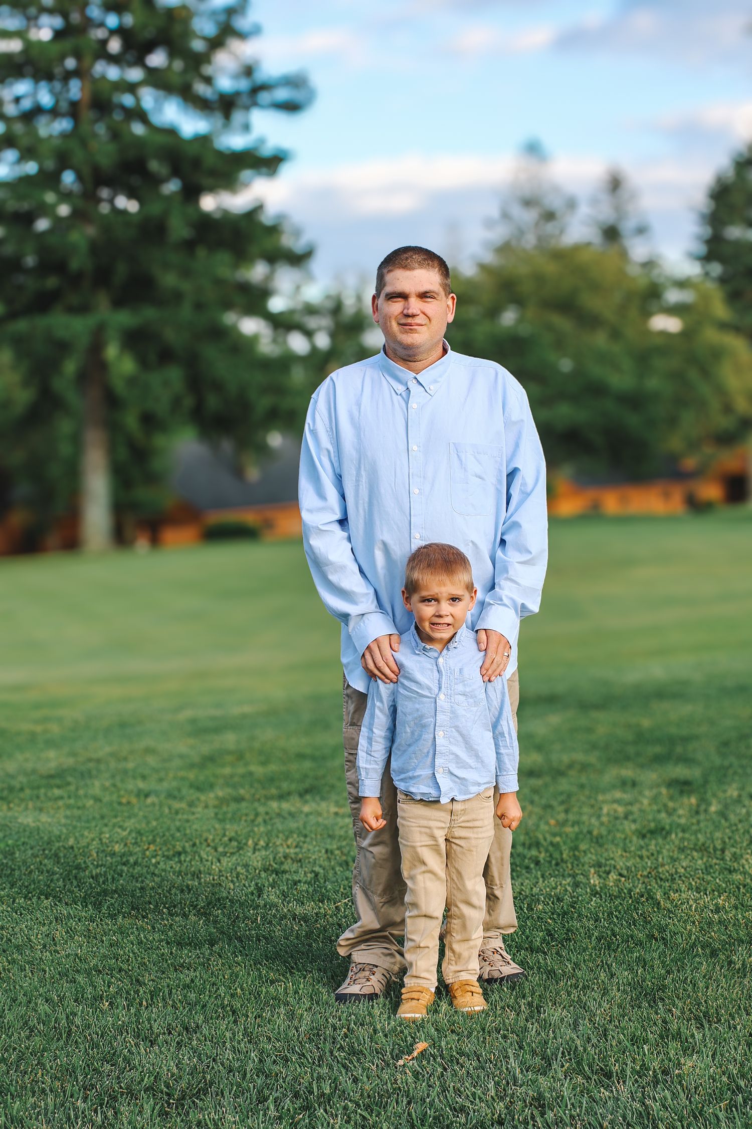 A parent and child stand together wearing matching light blue shirts and khaki pants on a grassy lawn.