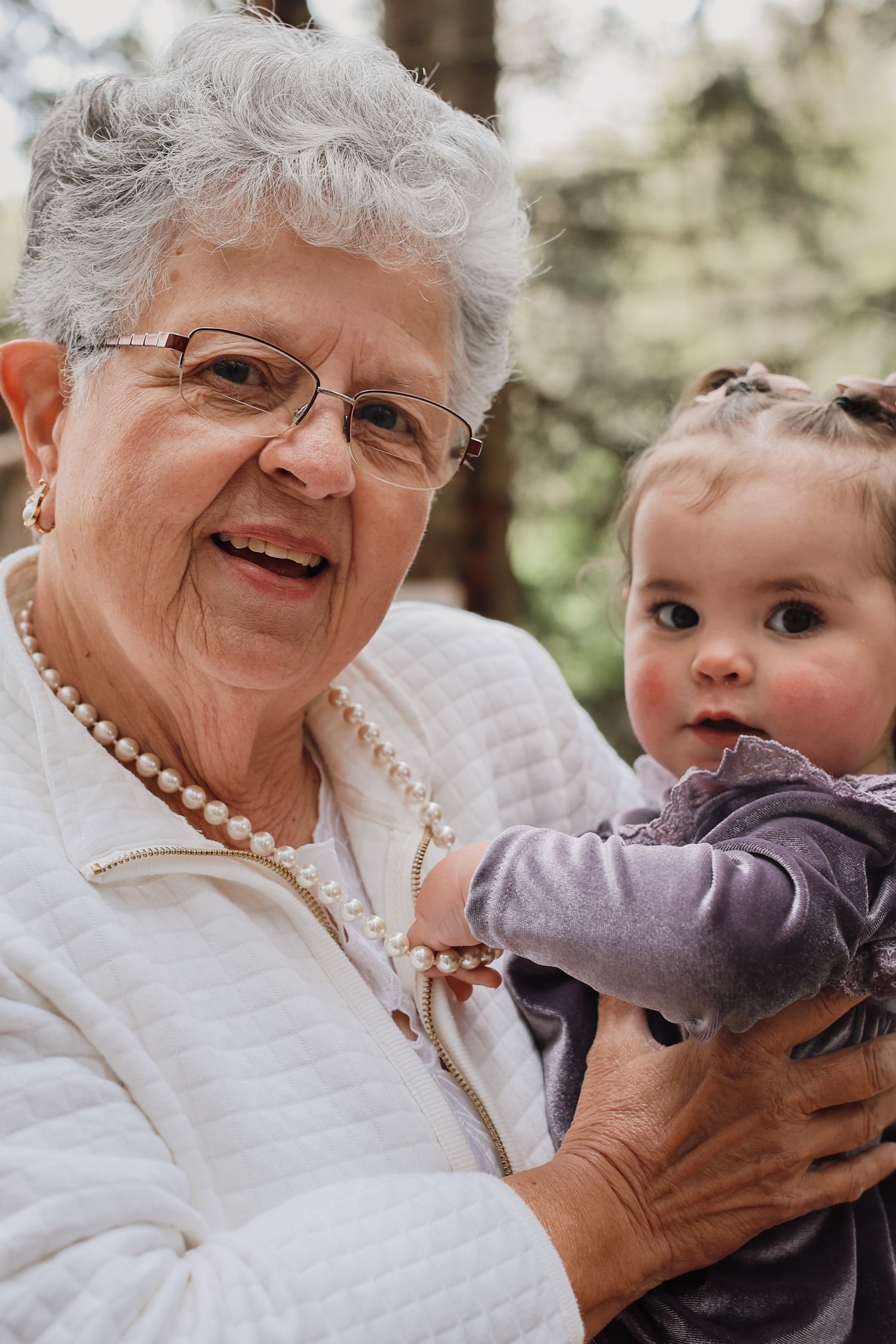 A heartwarming moment between an elderly woman in a white sweater and pearl necklace holding a small child outdoors.