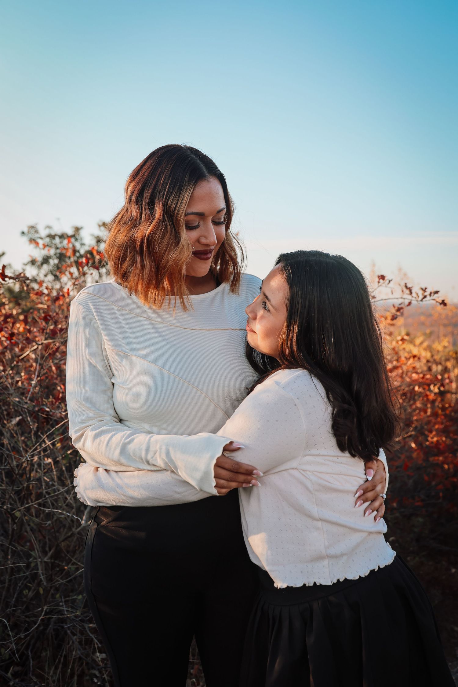 Two people in white sweaters embrace while sharing a tender moment at sunset among autumn foliage.