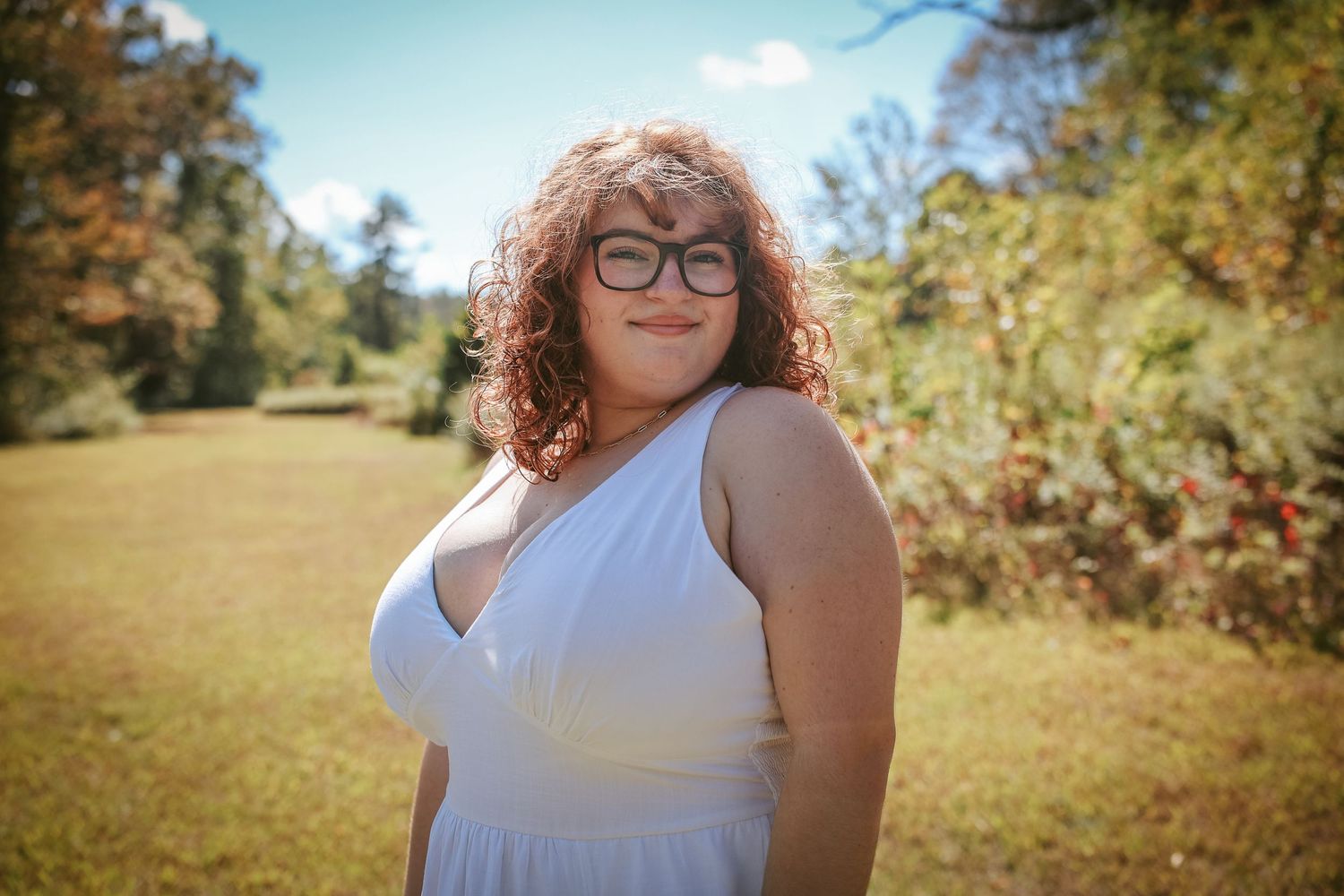 A person in a white sleeveless top poses outdoors in a sunny field with trees and natural scenery in the background.