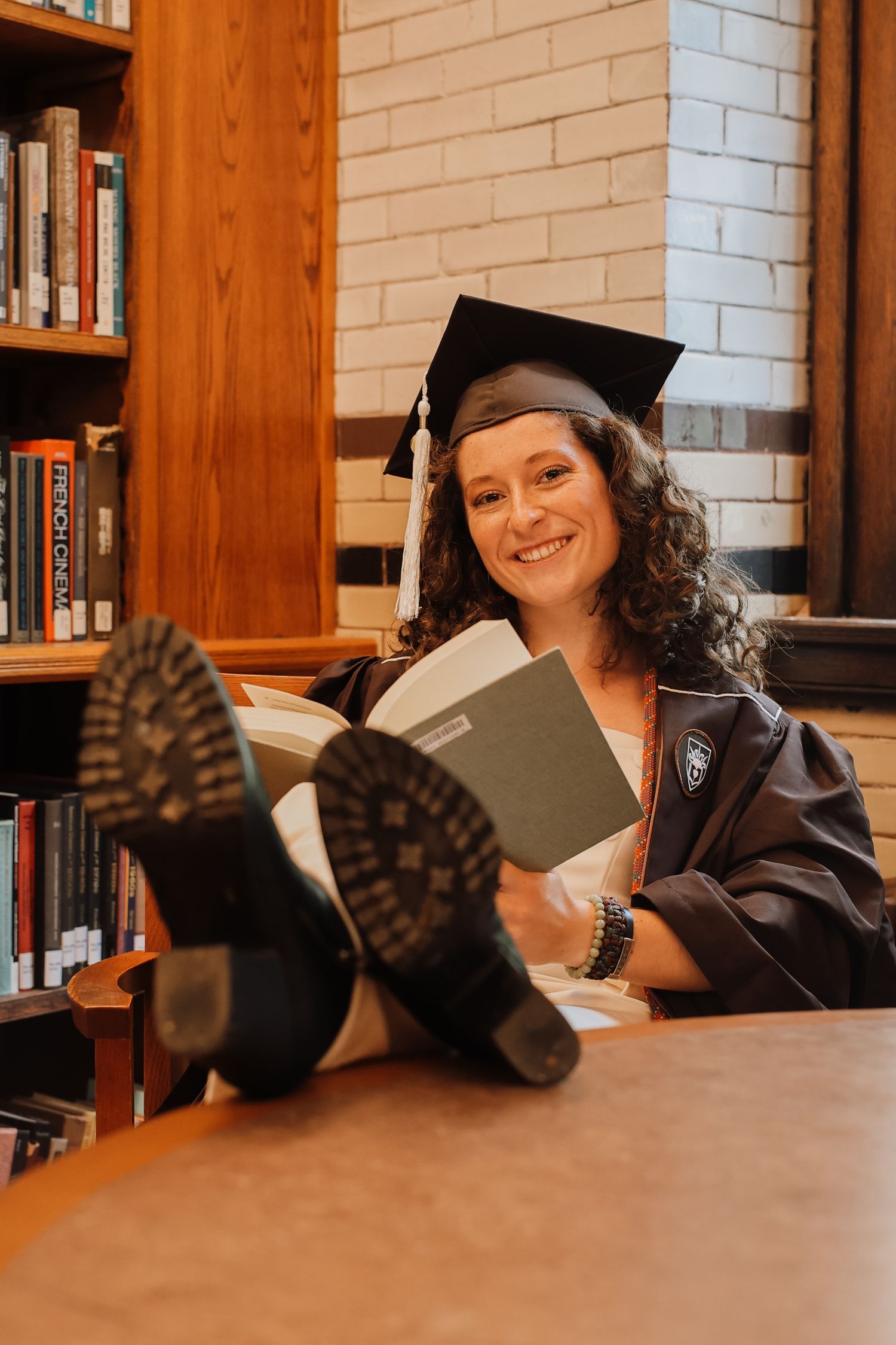 A graduate in cap and gown relaxes with feet up while reading in a library with brick walls.