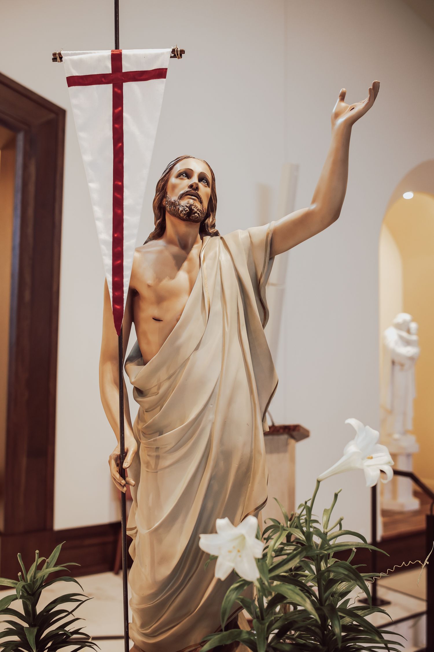 A sequence of religious statues in white robes with raised arms stand beside white lilies in an interior setting.