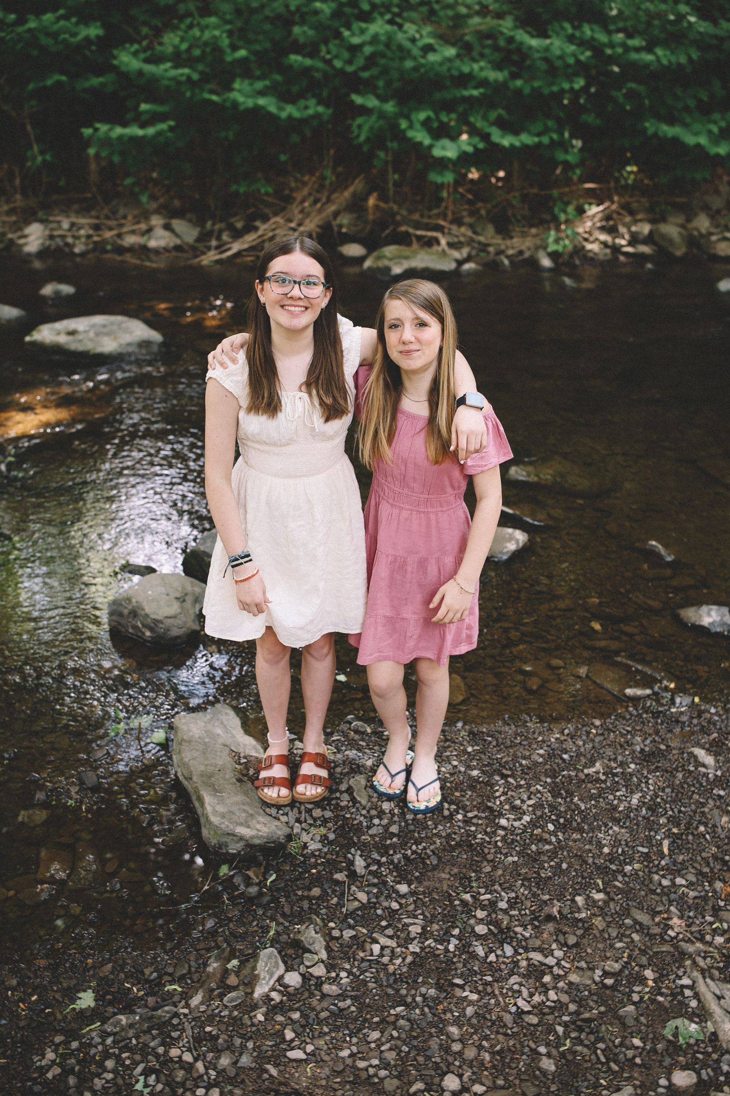 Two young friends sit together by a rocky creek bed surrounded by lush green forest scenery.