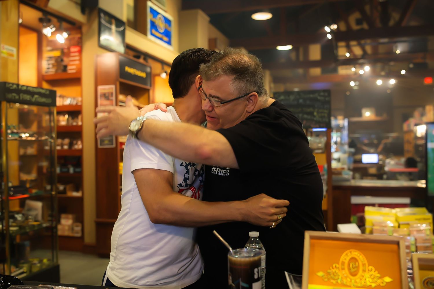 Two people embrace in a warm hug at a casual restaurant or bar setting.