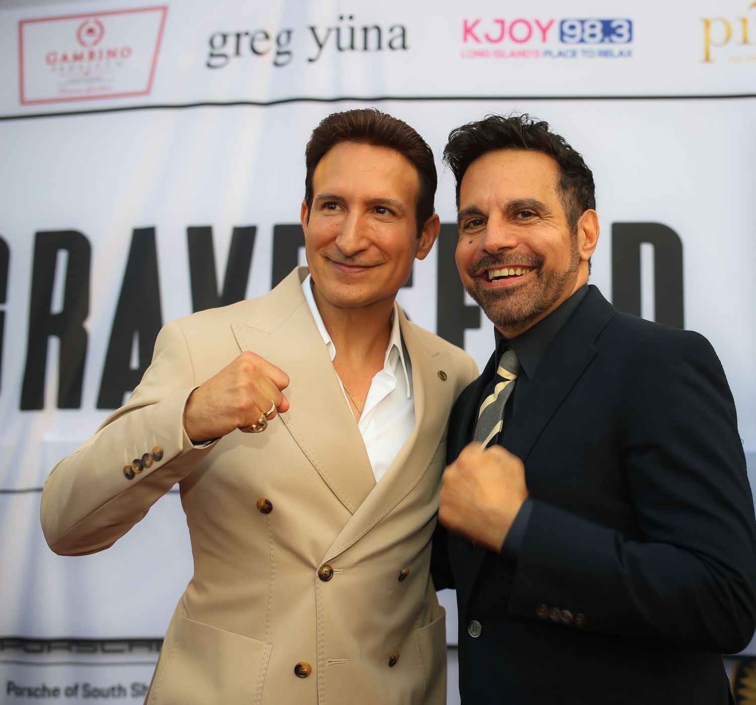 Two people in formal attire pose together at an event, one wearing a tan suit and the other in dark clothing.