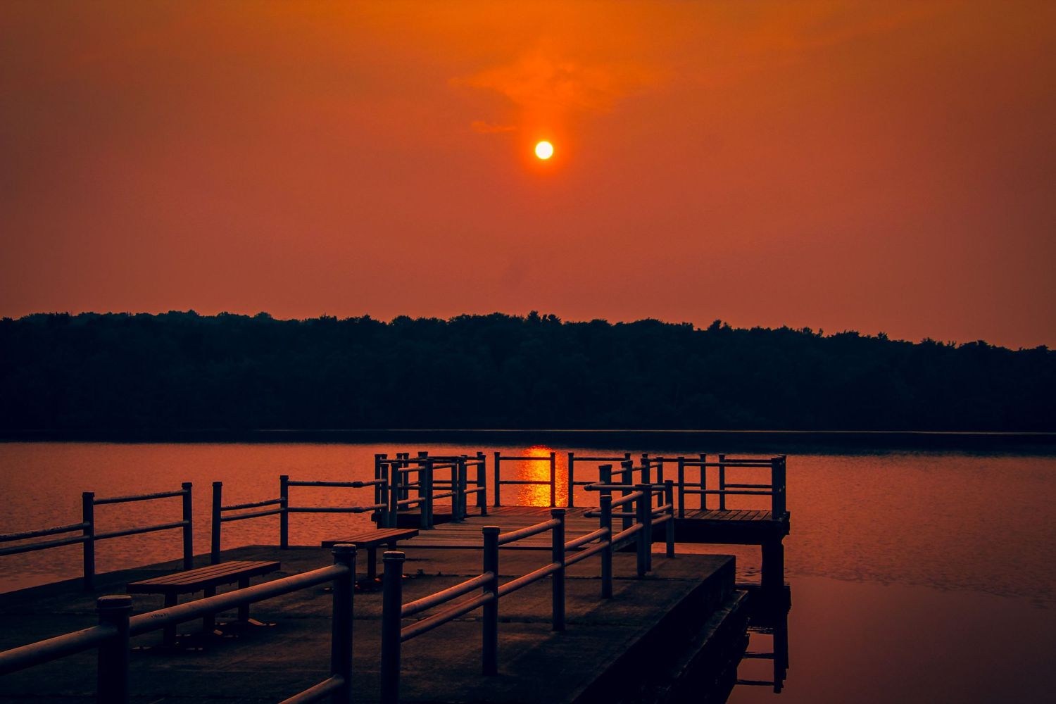 A wooden dock extends over calm waters at sunset with an orange sun glowing through a hazy sky above tree-lined shores.