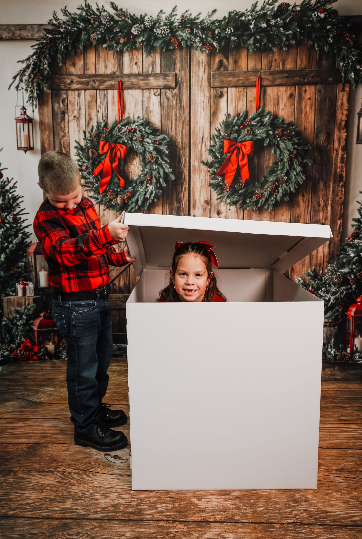 Holiday photo setup with rustic wood backdrop, evergreen garland, red bows and white display board on wooden floor.