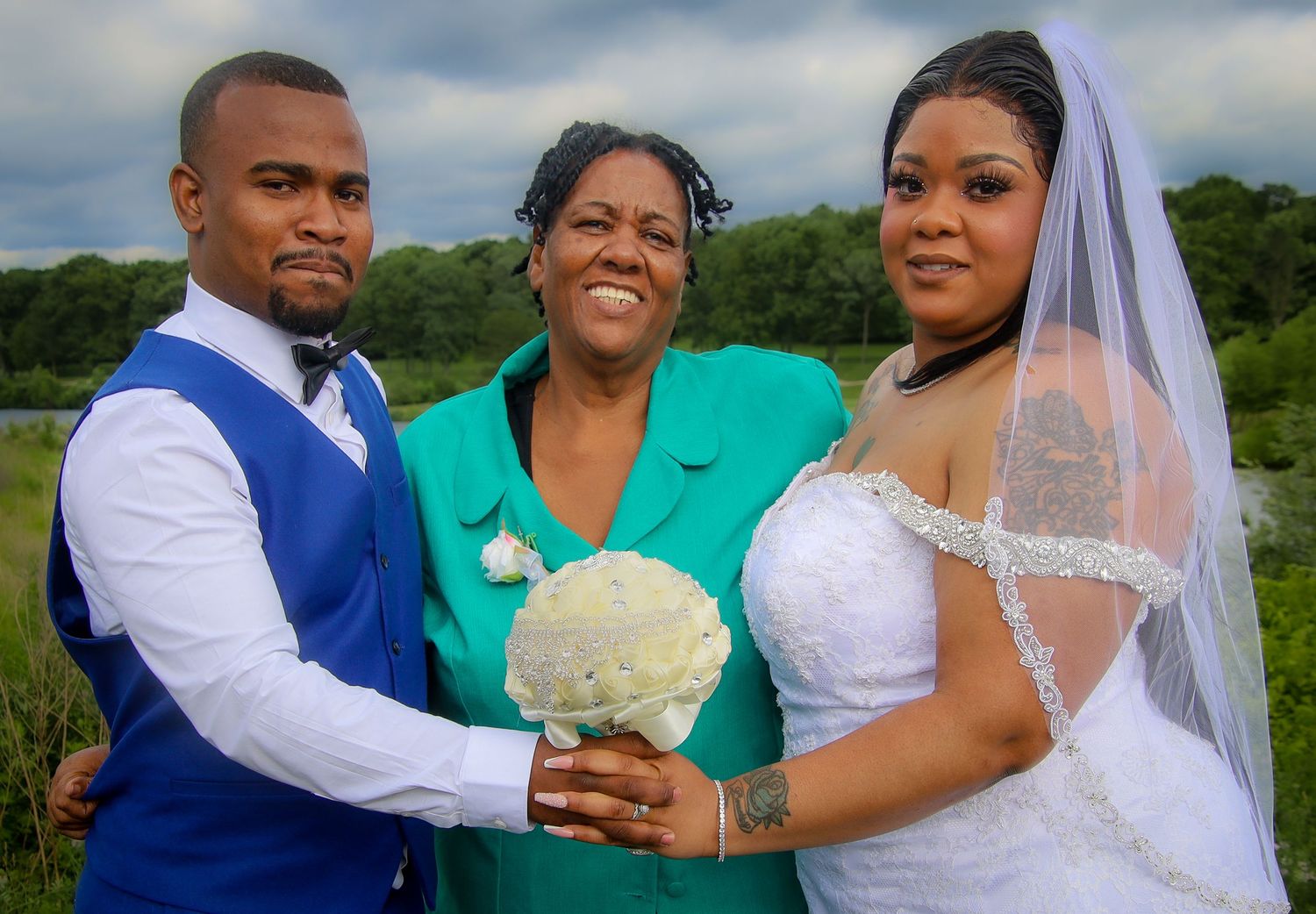 A joyful wedding photo outdoors with a bride in white, groom in blue vest, and guest in turquoise holding a bouquet.