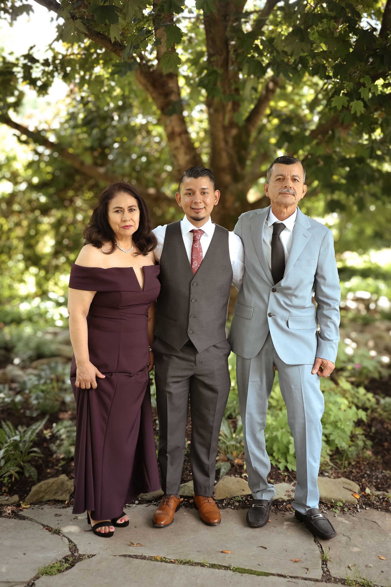 Three people in formal attire pose together outdoors in a garden setting with natural lighting.