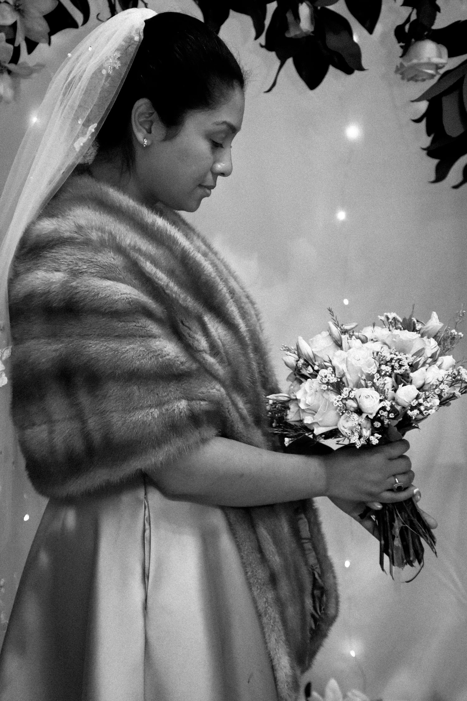 A bride in a white gown and fur stole holds a bouquet of flowers in a classic black and white photograph.