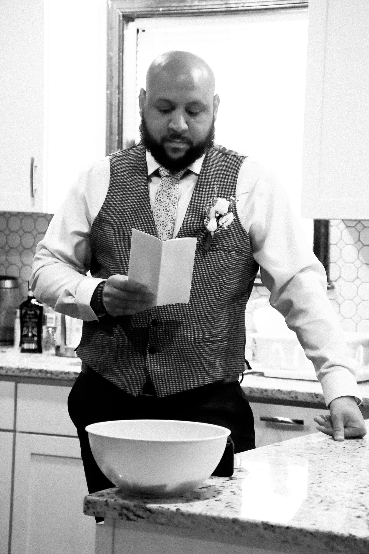 A person in a vest and tie reads from a card while standing in a kitchen area.