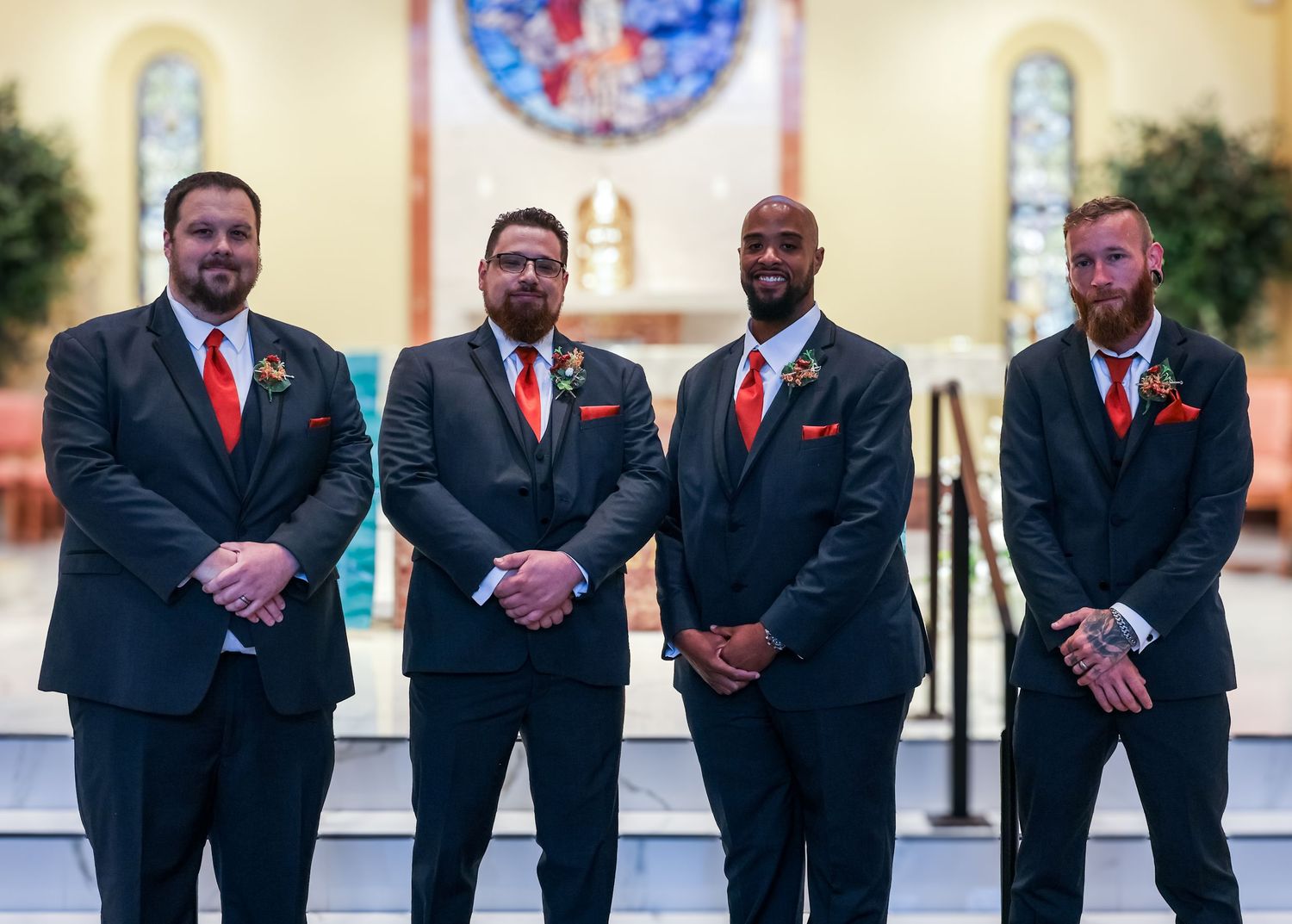 Wedding party members in black suits and red ties pose together outdoors at a formal event.