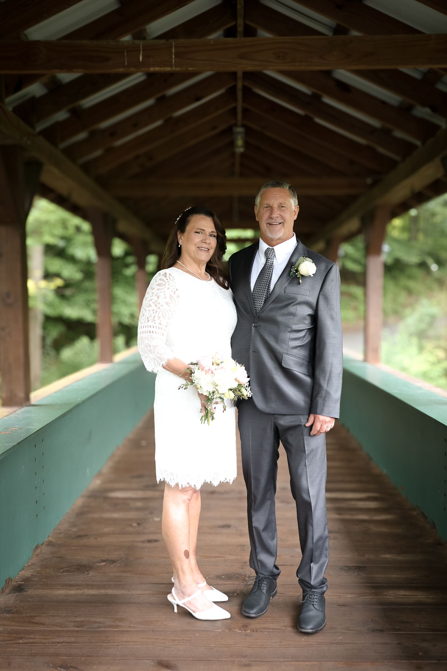 A couple poses together on a covered wooden bridge, the bride in a white lace dress and groom in a gray suit.