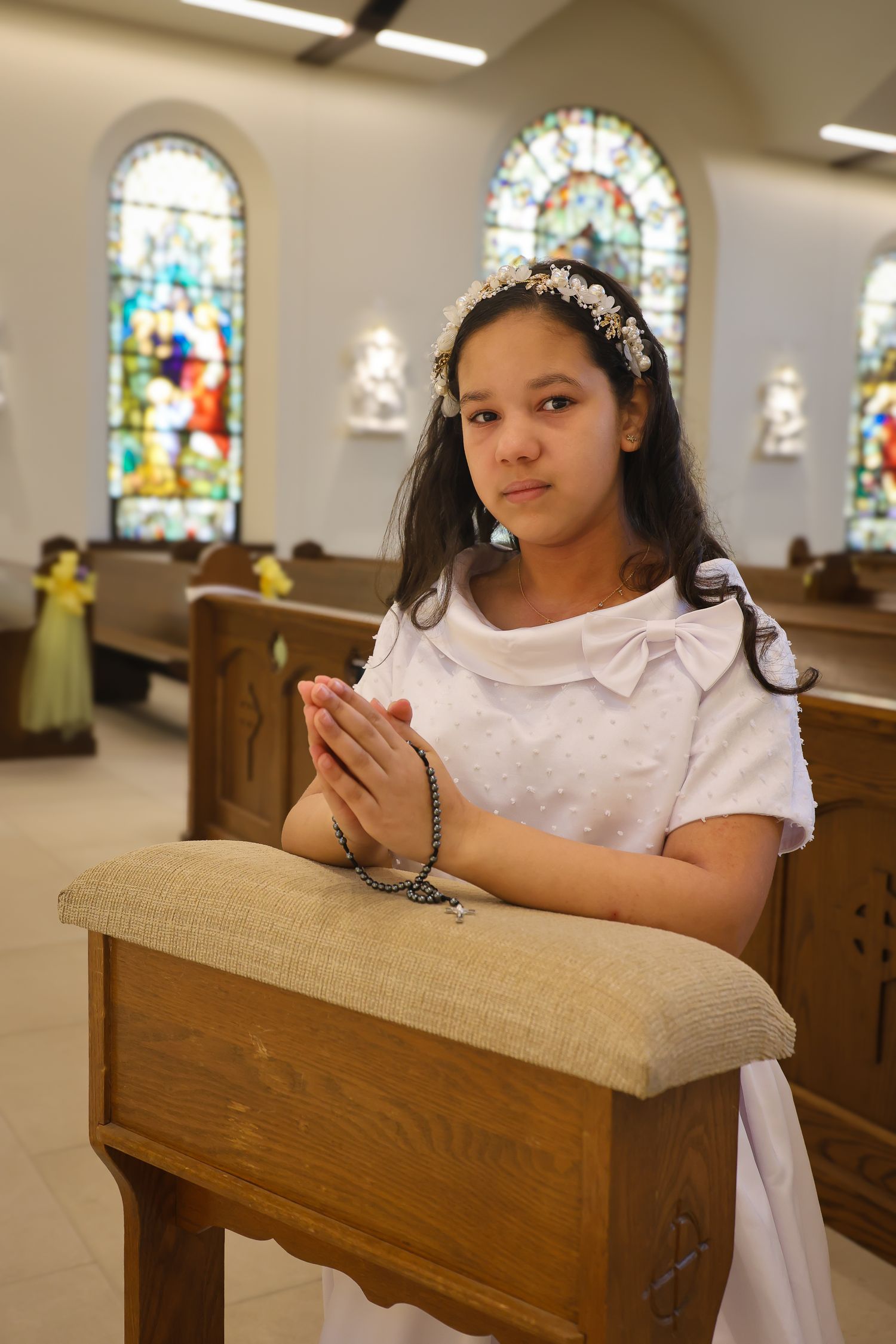 A child in white dress sits thoughtfully in a church pew with stained glass windows visible in the background.