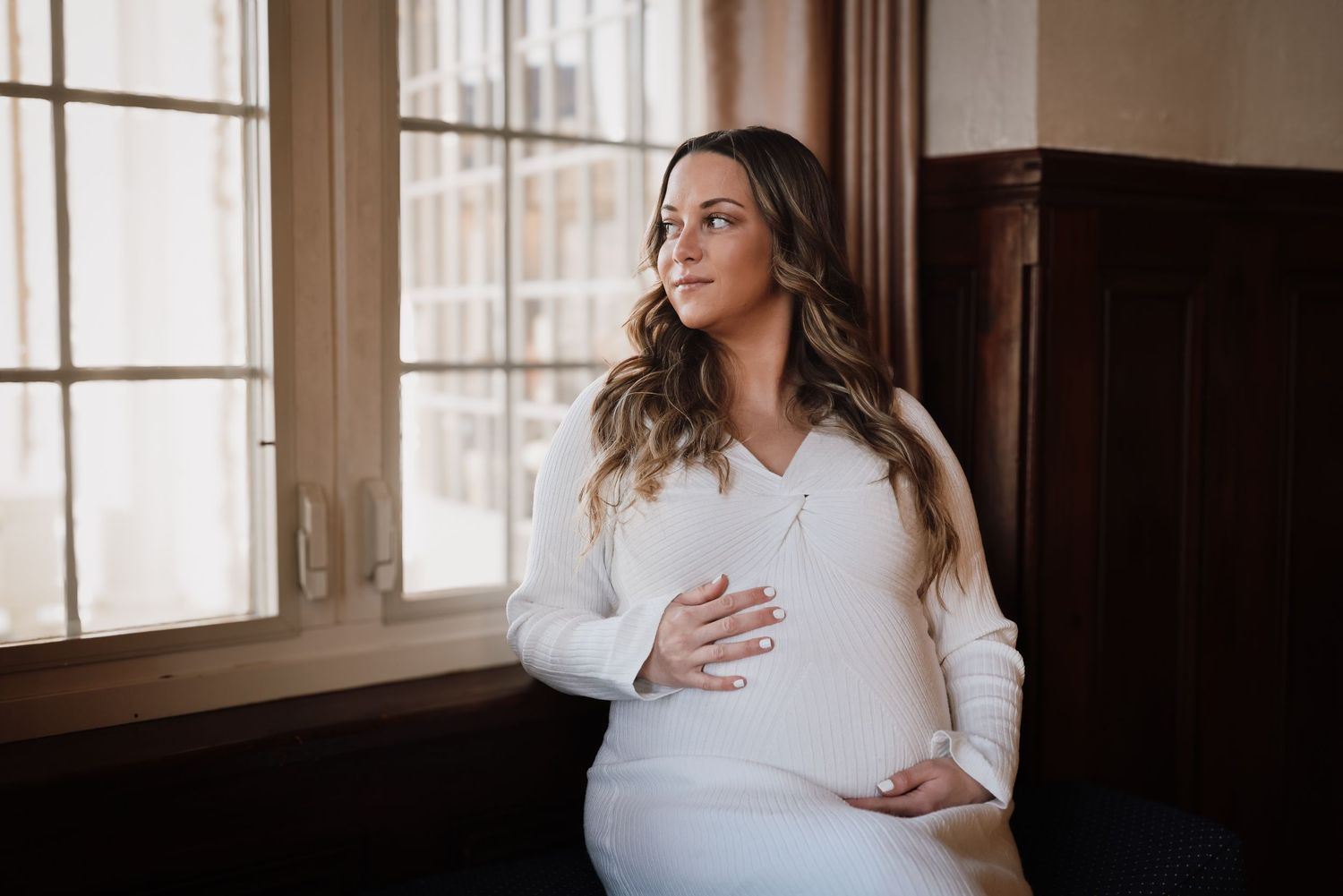 A woman in a white sweater sits by a window in natural light creating a cozy maternity portrait.