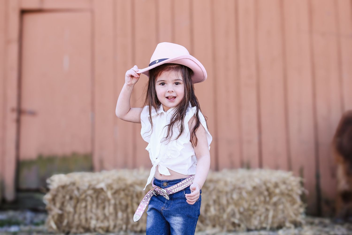 A young child in a pink cowboy hat and jeans poses playfully in front of hay bales on a farm.