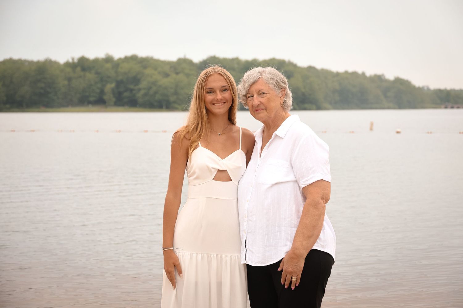 Two people in white outfits pose together by a serene lake with trees in the background during golden hour.